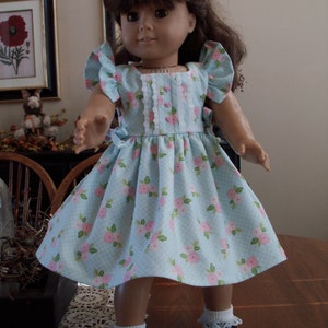 May include: A doll wearing a light blue dress with a pink floral pattern and white lace trim. The dress has puffed sleeves and a gathered skirt. The doll is wearing white socks and black shoes.