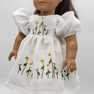 May include: A doll wearing a white dress with daisy embroidery. The dress has short, puffy sleeves and a lace trim at the hem. The doll has brown hair and is wearing black shoes with white socks.