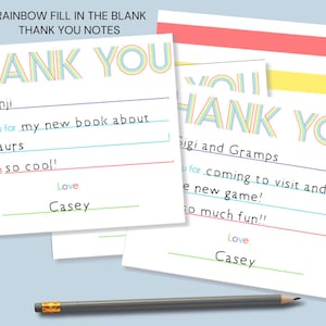 May include: A set of white fill-in-the-blank thank you notes featuring the text THANK YOU in rainbow-colored striped letters. The cards include lines for a recipient name, a message, and a sender name, accompanied by a gray wooden pencil.
