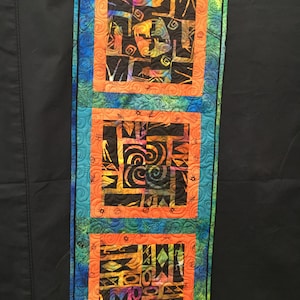 May include: A quilted table runner with three orange and black patterned squares framed by a teal border. The squares feature abstract designs with swirls and geometric shapes.