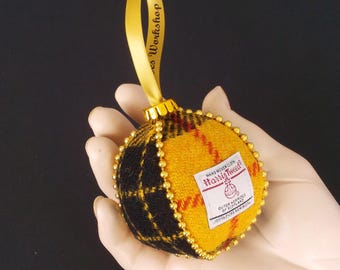 Puede incluir: Una bola de Navidad redonda con un diseño a cuadros amarillo y negro. El adorno está adornado con cuentas doradas y tiene una cinta amarilla para colgar. La etiqueta dice "Harris Tweed". La cinta dice "es Workshop".