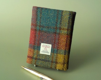 Puede incluir: Una funda rectangular para cuaderno hecha de lana Harris Tweed multicolor con un patrón de cuadros en azul verdoso, rojo y amarillo. Una etiqueta rectangular blanca está cosida en la parte delantera con el texto Harris Tweed, Outer Hebrides of Scotland, 100% Pure New Wool. Un bolígrafo dorado se encuentra delante.