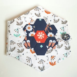 May include: A white fabric hexagon-shaped pouch with a floral print and a navy blue and orange flower-shaped applique. The pouch has a button closure.