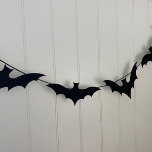 May include: A black bat garland with five bats hanging from a string. The bats are cut out of black paper and are hanging against a white wall.
