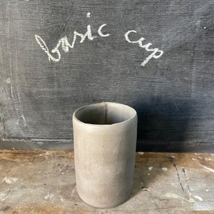 May include: A cylindrical, light gray cup with a slightly textured surface. The words "basic cup" are written in white chalk on a dark gray background behind the cup. The cup is sitting on a wooden surface.