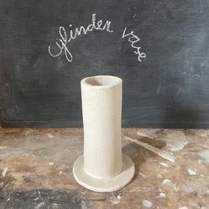 Könnte beinhalten: Eine handgefertigte, cremefarbene Keramik-Zylindervase mit strukturierter Oberfläche. Die Vase hat einen zylindrischen Körper und einen kreisförmigen Sockel. Die Worte "cylinder vase" sind mit weißer Kreide auf dunklem Hintergrund geschrieben.