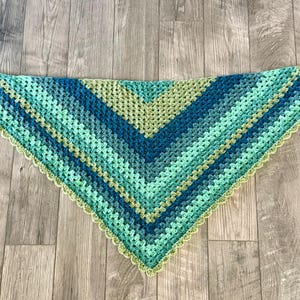 May include: A handmade crochet shawl in a triangular shape. The shawl features a gradient of blues and greens, with a scalloped edge. The color scheme includes shades of teal, turquoise, and a lighter green. The shawl is displayed on a wooden surface.