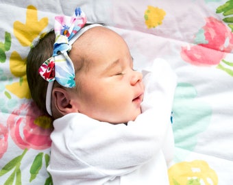 May include: A sleeping baby wearing a white onesie and a colorful floral headband on a white and pink floral sheet.