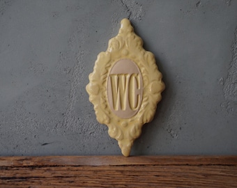 May include: A yellow ceramic sign with an ornate frame and the letters "WC" in a white oval in the center.