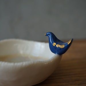 Può includere: Una ciotola in ceramica bianca con un manico a forma di uccello blu. L'uccello ha accenti dorati sulle ali.