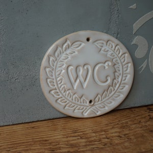 May include: White ceramic sign with a wreath of leaves around the letters "WC".