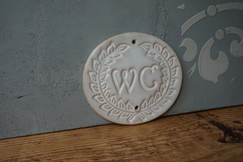 May include: White ceramic sign with a wreath of leaves around the letters "WC".