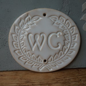 May include: White ceramic round sign with a wreath of leaves around the letters "WC".
