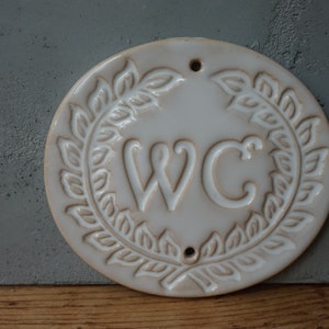 May include: White ceramic sign with a wreath of leaves around the letters "WC".