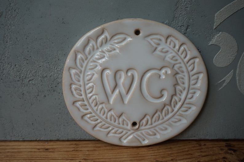 May include: White ceramic sign with a wreath of leaves around the letters "WC".