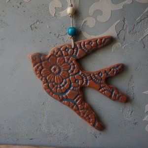 May include: A ceramic bird-shaped ornament in shades of brown and blue, suspended by a string with a wooden bead and a blue bead. The bird features a detailed floral pattern and is set against a textured gray background.