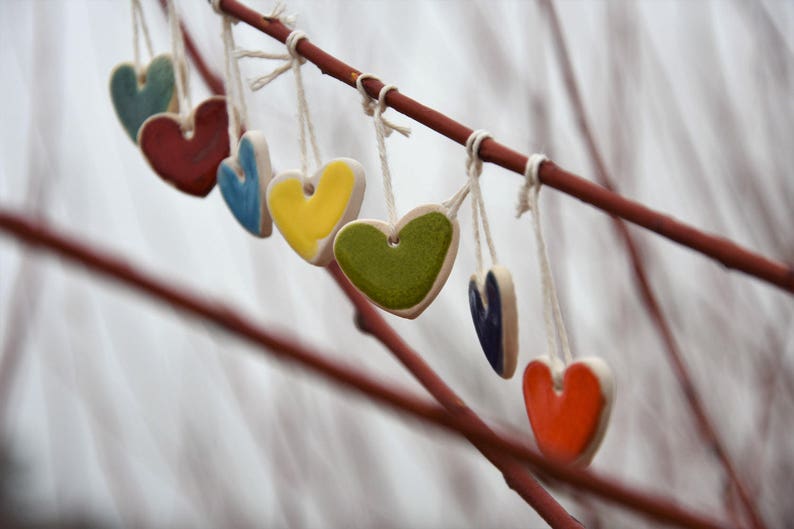 May include: A branch with seven ceramic heart ornaments hanging from it. The hearts are in various colors, including red, blue, yellow, green, and teal.