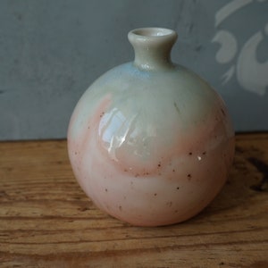 K&ouml;nnte beinhalten: Eine kleine, runde Keramikvase mit einer hellgr&uuml;nen und rosafarbenen Glasur. Die Vase hat eine leicht strukturierte Oberfl&auml;che und eine kleine &Ouml;ffnung oben.
