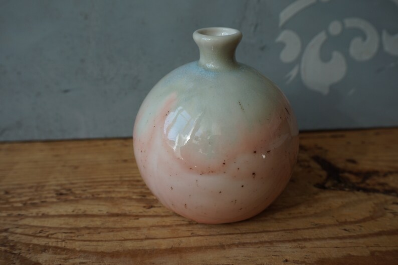 K&ouml;nnte beinhalten: Eine kleine, runde Keramikvase mit einer hellgr&uuml;nen und rosafarbenen Glasur. Die Vase hat eine leicht strukturierte Oberfl&auml;che und eine kleine &Ouml;ffnung oben.