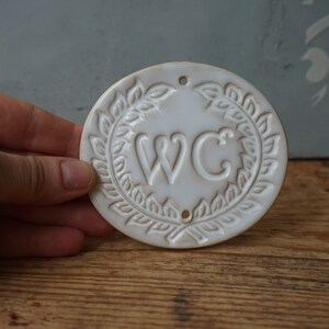 May include: White ceramic sign with a wreath design and the letters "WC" in the center.