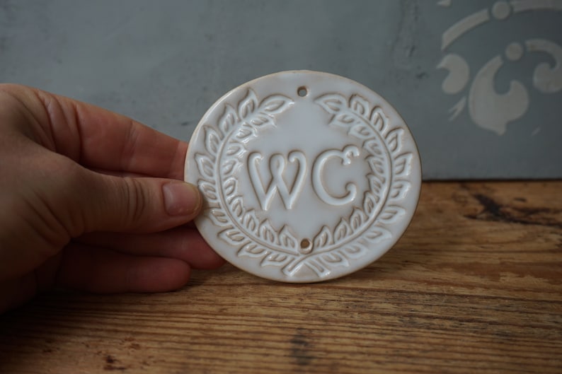 May include: White ceramic sign with a wreath design and the letters "WC" in the center.