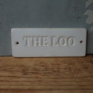 Könnte beinhalten: Ein rechteckiges, cremefarbenes Keramikschild mit den Worten "THE LOO" in Großbuchstaben geprägt. Das Schild hat zwei kleine Löcher zur Befestigung. Das Schild steht auf einer Holzoberfläche und einer grauen Wand.
