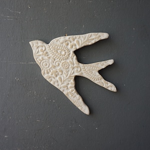 May include: A white ceramic bird with a detailed floral pattern. The bird is in flight with its wings spread out.
