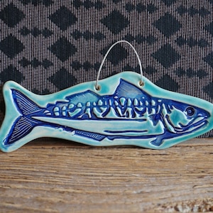 Könnte beinhalten: Ein blauer und grüner Keramikfisch-Anhänger mit einer Aufhängeschlaufe. Der Fisch hat ein detailliertes Design und eine strukturierte Oberfläche.