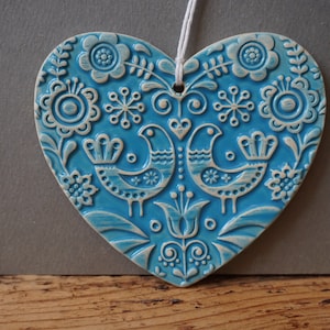 Op de afbeelding: Een blauwe, hartvormige keramische hanger met een wit bloemen- en vogelmotief. De hanger heeft een gat aan de bovenkant om op te hangen.