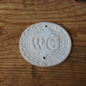 May include: White ceramic sign with a wreath design and the letters "WC" in the center.