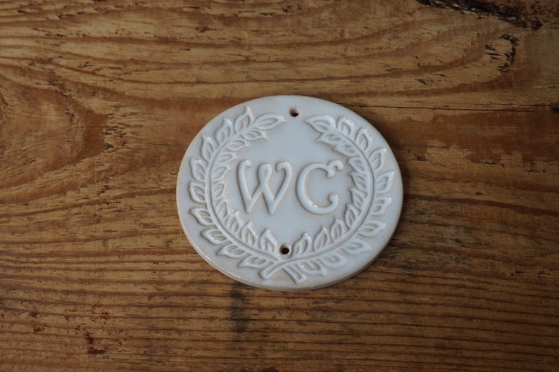 May include: White ceramic sign with a wreath design and the letters "WC" in the center.