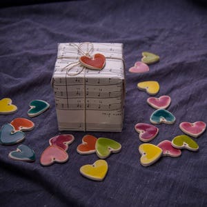 May include: A gift box wrapped in white paper with musical notes printed on it. A red heart charm is attached to the box with twine. The box is surrounded by colorful ceramic hearts on a blue fabric surface.