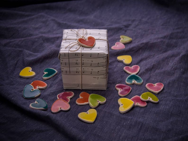May include: A gift box wrapped in white paper with musical notes printed on it. A red heart charm is attached to the box with twine. The box is surrounded by colorful ceramic hearts on a blue fabric surface.