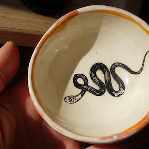 May include: A white ceramic bowl with a red and orange rim. The bowl has a black and white illustration of a snake on the inside.