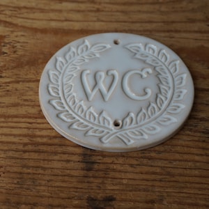 May include: White ceramic round sign with a wreath of leaves around the letters "WC".