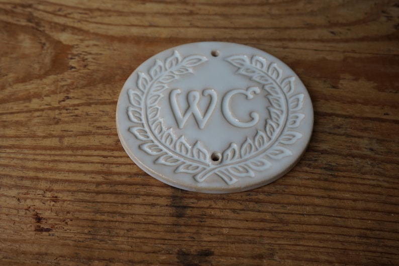 May include: White ceramic round sign with a wreath of leaves around the letters "WC".