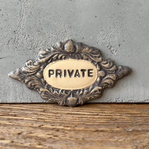 Puede incluir: Un letrero de metal decorativo con un diseño ornamentado y la palabra "PRIVATE" en letras doradas.