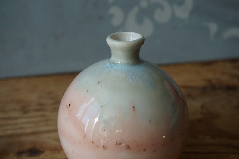 K&ouml;nnte beinhalten: Eine kleine, runde Keramikvase mit hellblauer und rosafarbener Glasur. Die Vase hat eine breite &Ouml;ffnung und einen schmalen Hals.
