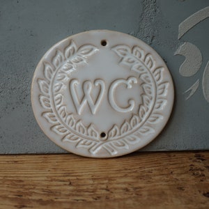 May include: White ceramic sign with a wreath of leaves around the letters "WC".