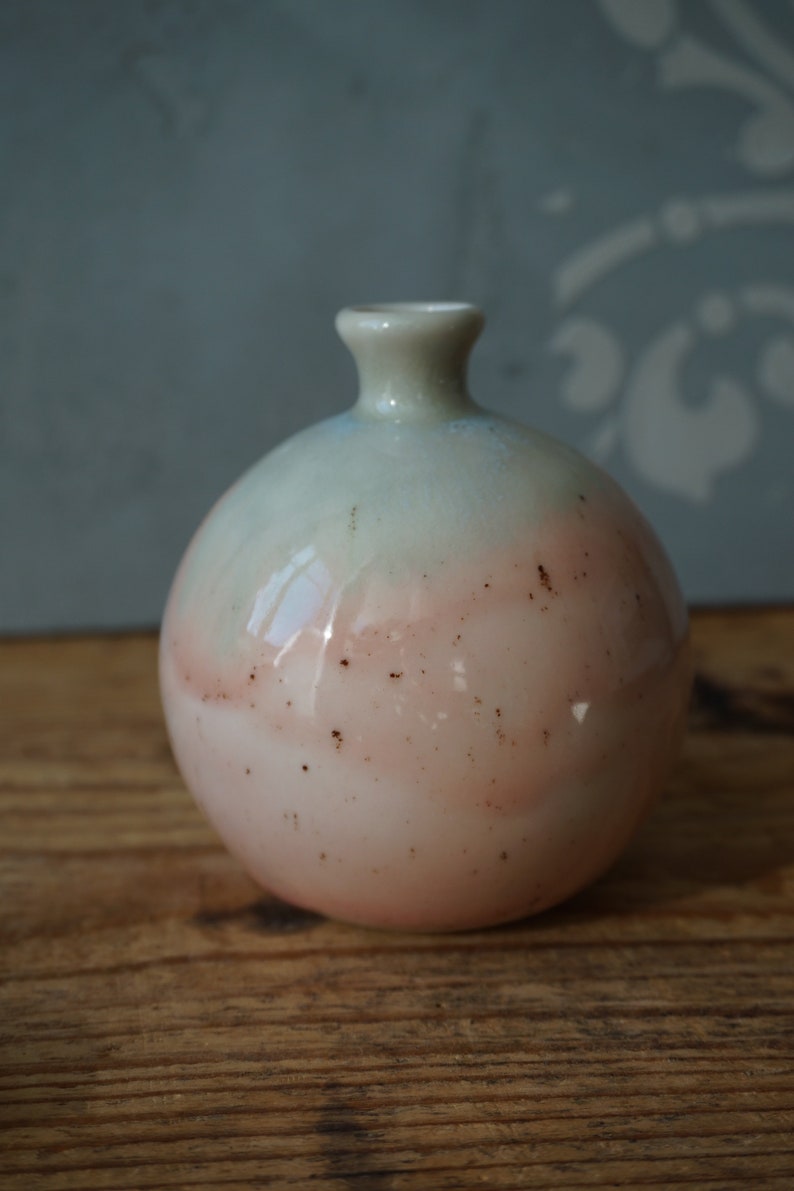K&ouml;nnte beinhalten: Eine kleine, runde Keramikvase mit hellblauem und rosafarbenem &Uuml;berzug. Die Vase hat einen gesprenkelten braunen &Uuml;berzug und eine leicht ausgestellte &Ouml;ffnung.