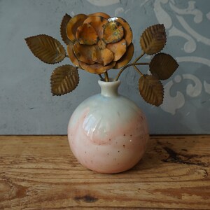K&ouml;nnte beinhalten: Eine kleine, runde, rosa-wei&szlig;e Keramikvase mit einer Metallrose und Bl&auml;ttern darin. Die Vase hat eine gesprenkelte Glasur.