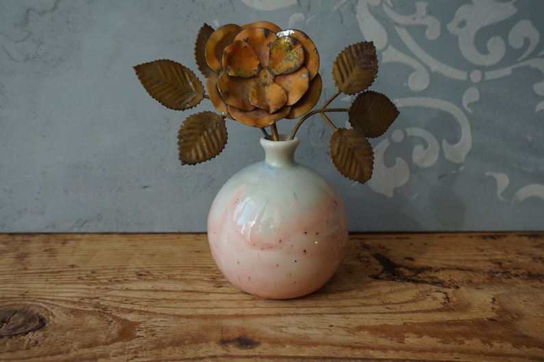 K&ouml;nnte beinhalten: Eine kleine, runde, rosa-wei&szlig;e Keramikvase mit einer Metallrose und Bl&auml;ttern darin. Die Vase hat eine gesprenkelte Glasur.