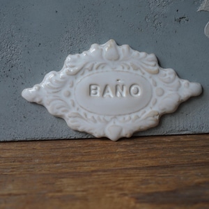 Op de afbeelding: Een wit keramisch bord met een sierlijk ontwerp en het woord "BAÑO" in het midden. Het bord is bevestigd aan een grijs oppervlak.