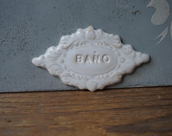 May include: White ceramic sign with an ornate design and the word "BAÑO" in the center. The sign is attached to a gray surface.