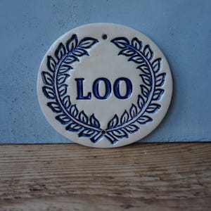 May include: White ceramic round sign with a blue wreath design and the word "LOO" in blue lettering.