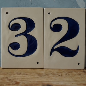 May include: Two rectangular ceramic house number tiles. The numbers 3 and 2 are in a dark blue, stylized font. The tiles are off-white with a glossy finish and have a small hole at the top for hanging. The background is a light blue.