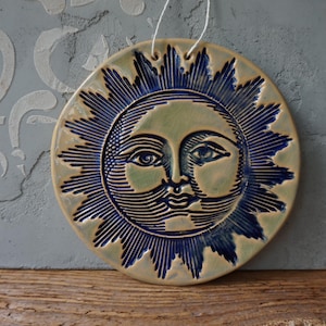 May include: A circular ceramic sun face wall hanging. The sun features a light green face with blue rays and details. It is suspended by a white string. The background is a light blue wall.