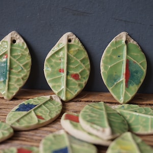 Op de afbeelding: Een verzameling keramische bladeren met groene glazuur en kleurrijke accenten. De bladeren hebben een getextureerd oppervlak en zijn perfect om een ​​natuurlijke touch toe te voegen aan sieraden of knutselwerk.