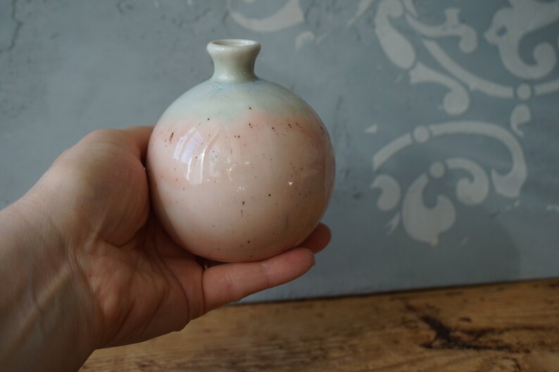 K&ouml;nnte beinhalten: Eine kleine, runde Keramikvase mit hellblauer und rosafarbener Glasur. Die Vase hat eine gesprenkelte Oberfl&auml;che und eine schmale &Ouml;ffnung oben.