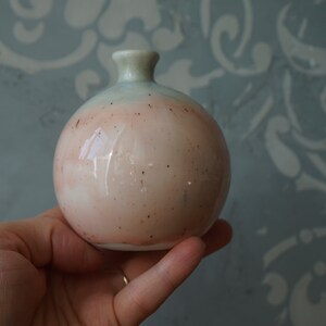 K&ouml;nnte beinhalten: Eine kleine, runde Keramikvase mit hellblauer und rosafarbener Glasur. Die Vase hat eine gesprenkelte Oberfl&auml;che und eine leicht abgerundete &Ouml;ffnung.